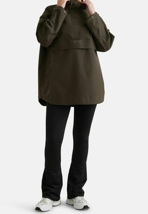 Person, der justerer kraven på en oversized olivengrøn pullover, iført sorte tætsiddende bukser og hvide sneakers, stående på hvid baggrund.