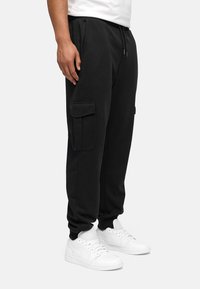 Pantalones de chándal negros con cintura ajustable, bolsillos laterales y un gran bolsillo cargo lateral. Tejido suave con un corte holgado, combinados con zapatillas blancas.