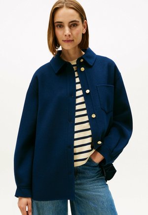 Navyblauwe wollen shirtjacket met een kraag, gouden knopen en een borstzak, gedragen over een gestreept shirt en gecombineerd met jeans.