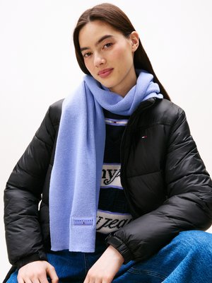 Écharpe tricotée bleu clair portée sur un pull à motifs et une veste en duvet noire. Étiquette notable avec le nom de la marque sur l'écharpe.