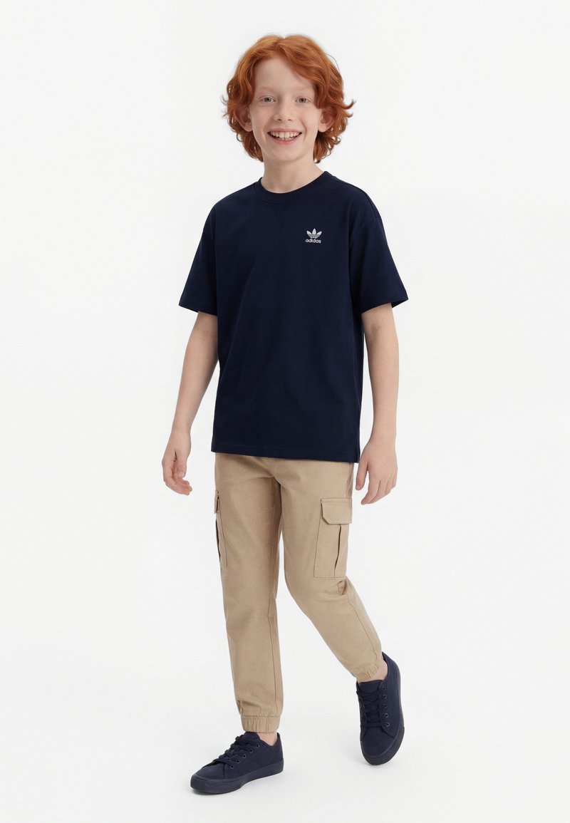 Naeratav punase juustega poiss kõnnib, seljas must Adidas T-särk, beežid tööpüksid ja mustad tossud, valgel taustal.