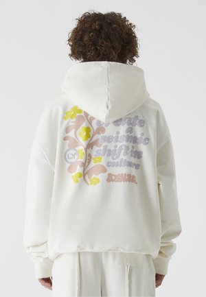 Personne aux cheveux bouclés porte un sweat à capuche écru avec un motif floral pastel et le texte « Créez un changement sismique dans la culture » au dos.