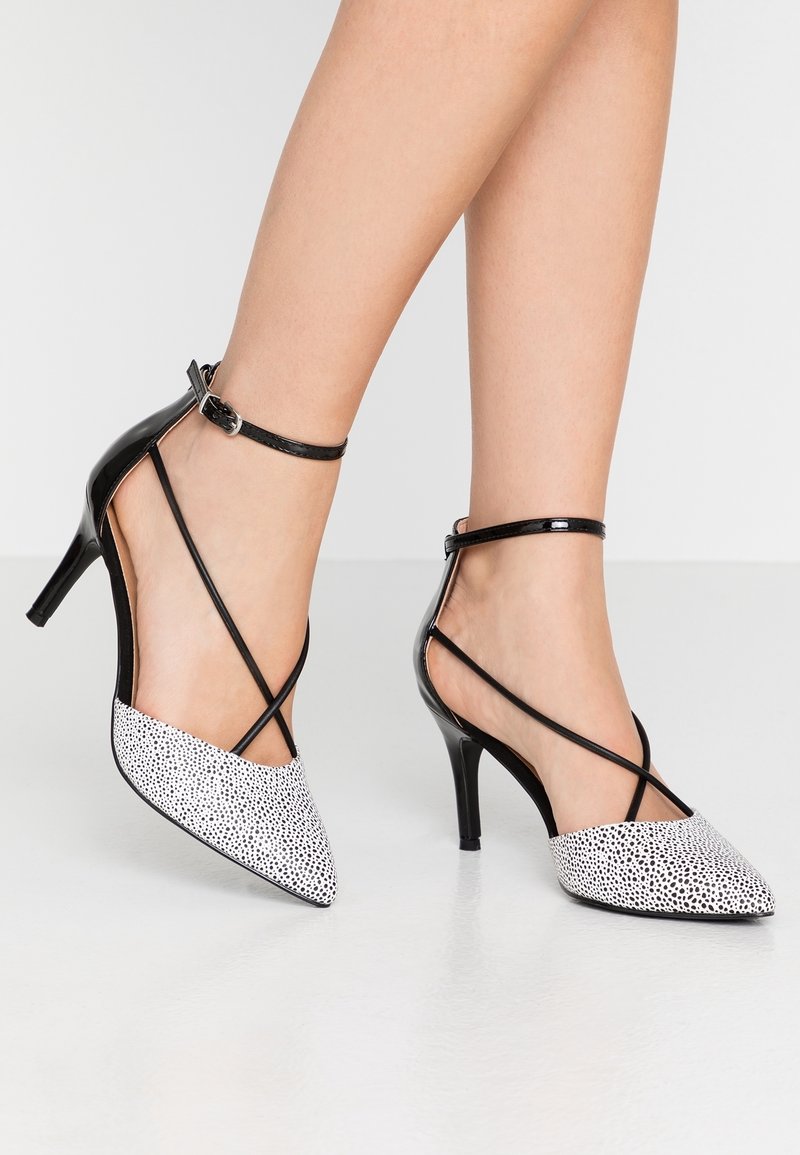 Escarpins à talons hauts pointus noirs et blancs à pois, avec de fines lanières autour de la cheville, portés sur des jambes croisées.