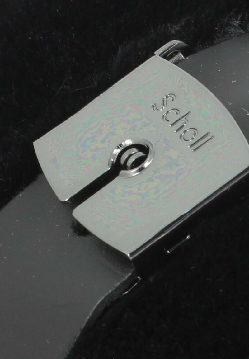 Schwarzes Lederband mit einer silbernen Metallschnalle, die mit "Scholl" graviert ist. Die Schnalle hat in der Mitte ein kreisförmiges Ausschnitt für die Befestigung.
