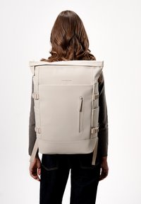 Beige rolovací batoh vyrobený z odolného materiálu. Disponuje nastavovacími popruhy, kapsou na zip a bočními sponami. Hladký texture, minimalistický design.