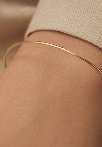 Gouden metalen bangle armband die rust op een blote arm; gladde oppervlak, dun ontwerp, iets reflecterende afwerking, minimalistische stijl.