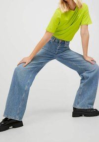 Person som bär en starkt grön T-shirt, vida blå jeans och svarta klumpiga skor, med benen utspridda och händerna vilande på låren.