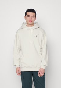 Felpa leggera beige con tasca a canguro, cappuccio con cordino e piccolo logo nero sul petto. Realizzata in un tessuto morbido e testurizzato.