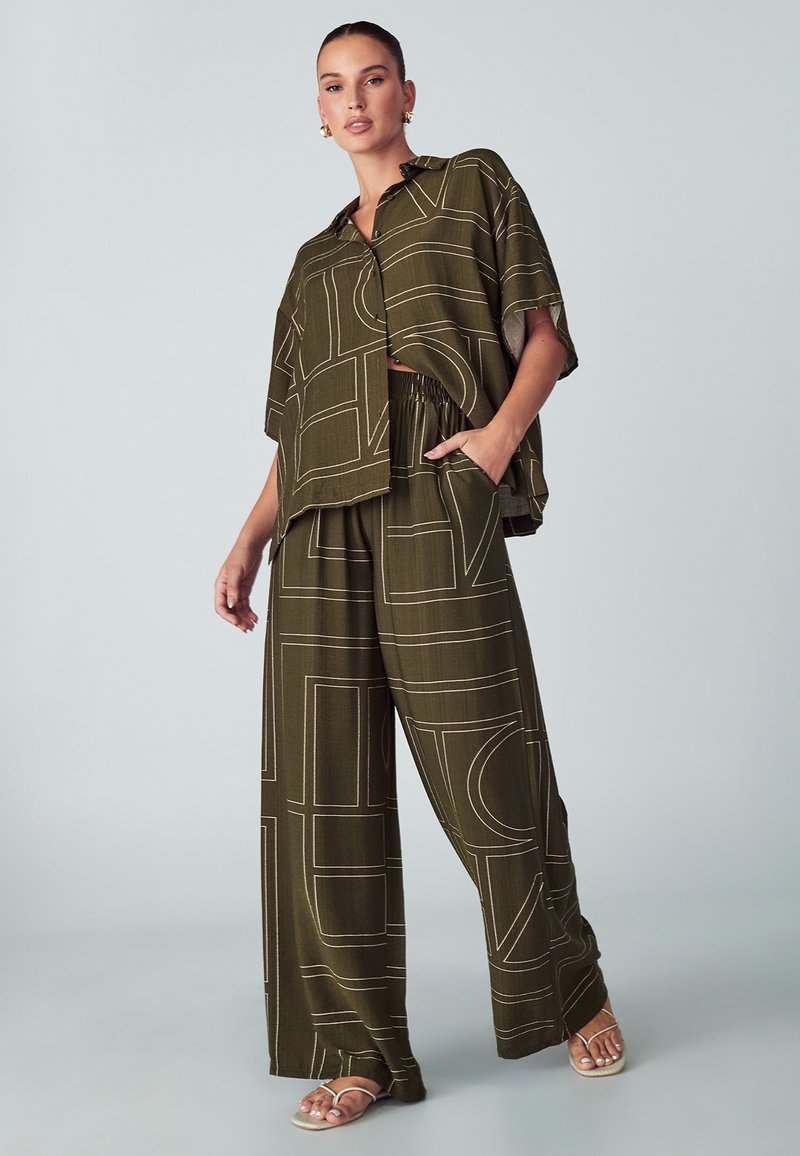Žena u maslinastozelenim širokim hlačama i odgovarajućoj slobodnoj košulji s kratkim rukavima, koja ima bijele geometrijske linije, stoji s jednom rukom u džepu.