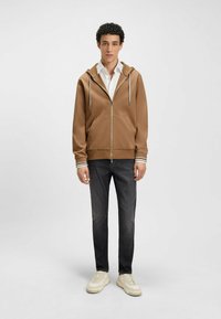 Brown zip-up hoodie with two front pockets, white drawstrings, and striped cuffs, paired with dark jeans and light sneakers.