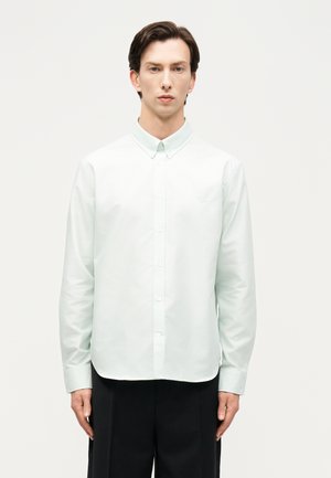 Άνδρας με κοντά σκούρα μαλλιά φορώντας ένα λευκό πουκάμισο με κουμπιά μακρυμάνικο και μαύρο παντελόνι μπροστά σε απλό ανοιχτό φόντο.