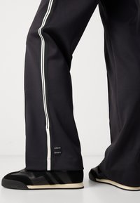 Pantalons de sport en mesh noirs avec des rayures latérales blanches et une étiquette de logo à l'ourlet, associés à des chaussures de sport noires et blanches présentant un design texturé.