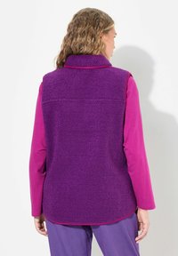 Lila Fleece-Weste mit hohem Kragen, strukturierter Oberfläche und seitlichen Schlitzen, getragen über einem pinken Langarmoberteil. Die Rückansicht zeigt die Länge des Kleidungsstücks.