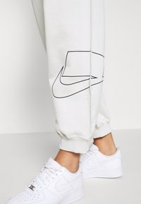 Ljusgrå sweatpants med en svart Nike-logotyptryck längs sidan, uppvikta anklar, i kombination med vita Nike Air Force 1 sneakers.