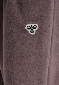 Zbliżenie na brązową tkaninę polaru z małym czarno-białym wyszywanym logo pszczoły umieszczonym na środku materiału.