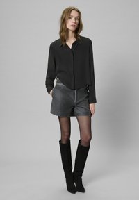 Kobieta w czarnej bluzie z długim rękawem, szarych skórzanych szortach, przeźroczystych czarnych rajstopach i czarnych kozakach do kolan stoi na szarym tle.