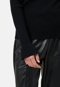 Czarny sweter z prążkowanym wzorem i rozszerzanymi mankietami, w połączeniu z czarnymi spodniami z sztucznej skóry. Tekstura jest gładka, podkreślająca elegancki, monochromatyczny wygląd.