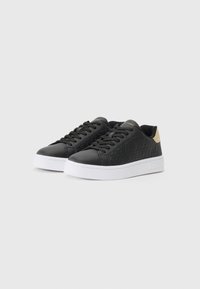 Svart sneakers med mønstrets syntetisk overdel, hvit gummisåle, flate snørebånd og en liten tan-farge aksent på hælen.