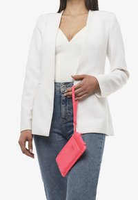Borsa compatta in pelle stampata coccodrillo rosa con tracolla, appesa alla mano di una persona. Indossata con un blazer bianco e jeans blu.