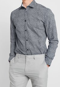 Långärmad skjorta med knappar i marinblått med vitt blommönster, med klassisk krage och uppvikta ärmar, kombinerad med ljusgrå byxor.