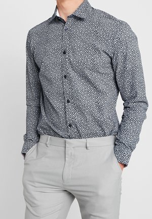 Camisa de botón de manga larga en azul marino con un patrón floral blanco, presentando un cuello clásico y mangas con puños, combinada con pantalones gris claro.