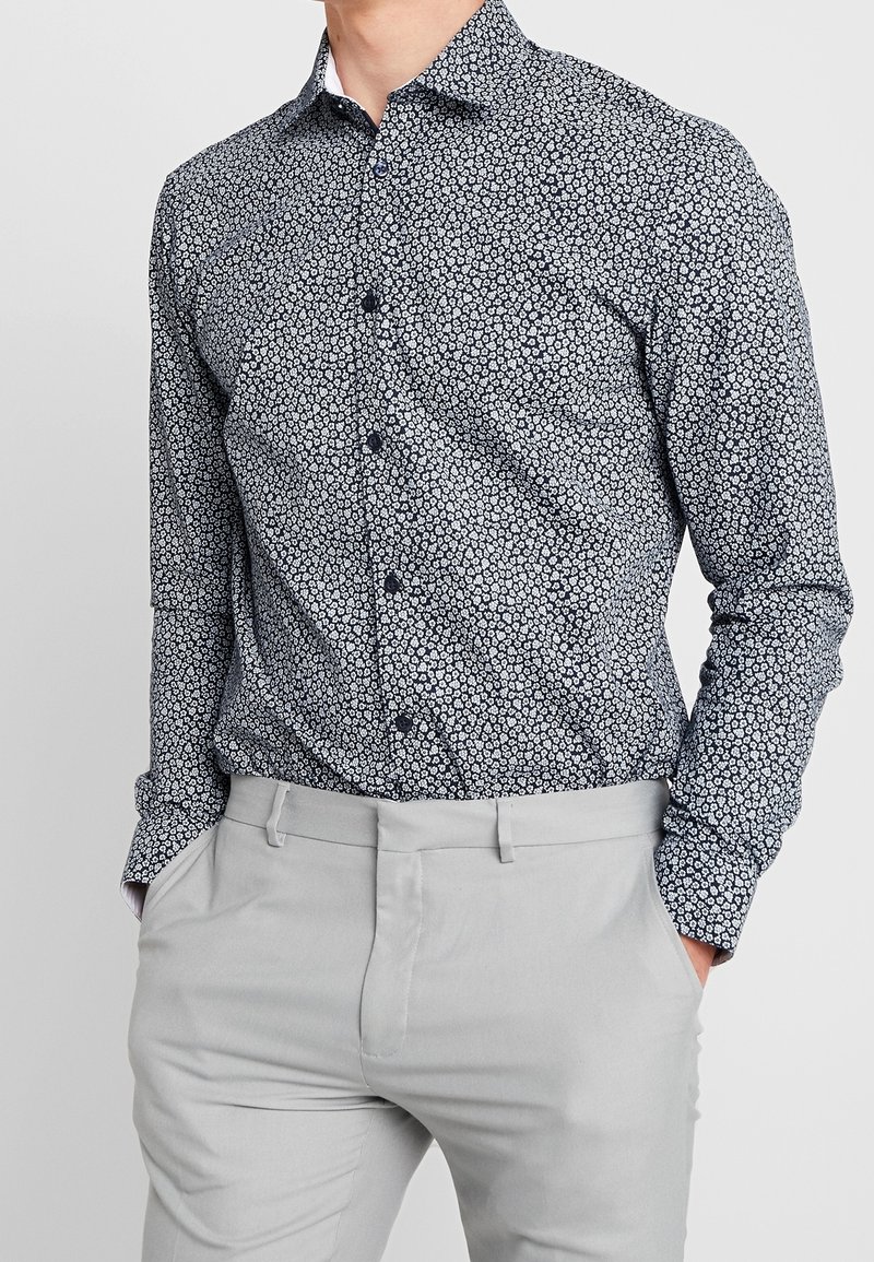 Långärmad skjorta med knappar i marinblått med vitt blommönster, med klassisk krage och uppvikta ärmar, kombinerad med ljusgrå byxor.