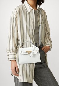Biała skórzana torebka o wyraźnej formie, z złotymi elementami i odpinanym paskiem, trzymana przez osobę w paskowanej koszuli i szarych dżinsach.