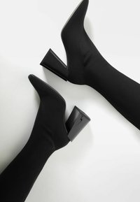 Botas negras hasta el tobillo con punta afilada y tejido texturizado. Tacones gruesos de charol negro ofrecen contraste. Textura suave, diseño elegante.