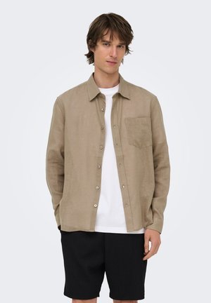 Jeune homme portant une chemise beige à boutons par-dessus un T-shirt blanc et un short noir, debout devant un fond clair uni.