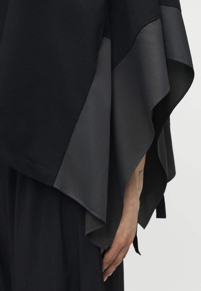 Main d'une personne posée à côté d'un tissu asymétrique superposé noir et gris foncé avec des bords fluides, un tatouage subtil étant visible sur l'avant-bras.