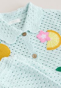 Jersey de bebé de ganchillo azul claro con botones de madera, cuello ondulado y parches de colores con una rodaja de limón, una flor rosa y una naranja.
