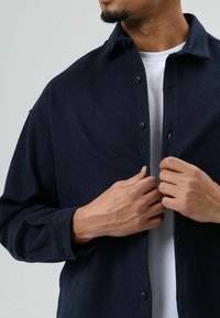 Donkerblauw overhemdbloes met knopen, kraag, gestructureerde stof en een ontspannen pasvorm. De lange mouwen zijn licht opgerold, waardoor een wit T-shirt eronder zichtbaar is.