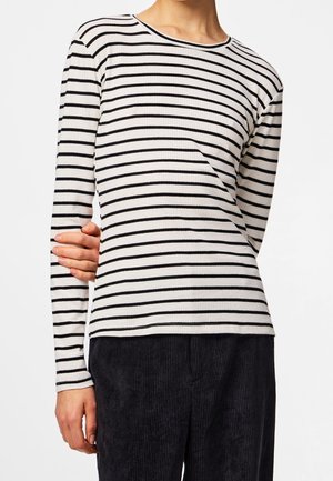 Personne portant une chemise blanche à manches longues avec des rayures horizontales noires et un pantalon en velours côtelé noir, main reposant sur la taille.