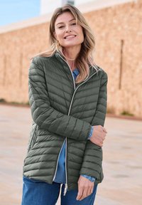 Gequilte, olijfgroene jas met een opstaand kraag, zilveren rits en een gladde textuur. Draagt over een blauw denim shirt.