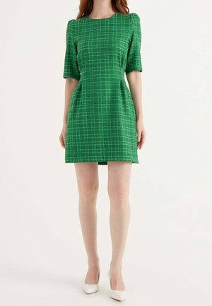 Nainen pukeutuneena lyhythihaiseen vihreään ruutukuosiseen mekkoon, jossa on pumpulihihat, ja valkoisiin korkokenkiin, seisoo yksinkertaisen taustan edessä.