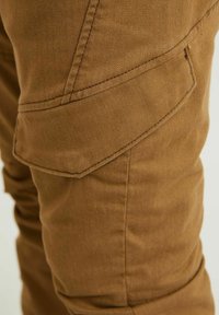 Gros plan sur la jambe d'un pantalon en tissu marron avec une poche à rabat inclinée et des détails de double surpiqûre visibles.