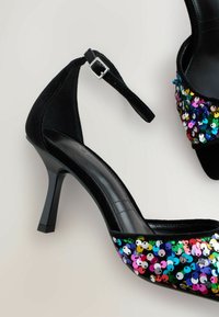 Scarpe con tacco alto nere con texture in velluto e un design di paillettes multicolori. Presentano una cinghia alla caviglia e un tacco quadrato.