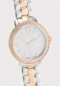 Montre-bracelet en argent et or rose avec un cadran argenté étincelant, une lunette ornée de cristaux et un design de bracelet en maillons. Fabriquée en Suisse.