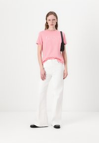 Rosa t-shirt med kort ärm och "GANT"-logotyp, i kombination med vita utsvängda byxor. Minimalistisk svart väska och platta skor med en sk shiny-accent.