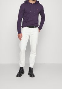 Felpa viola con cappuccio e tasca a marsupio; jeans slim-fit bianchi; cintura nera e stivali alla caviglia; texture liscia.