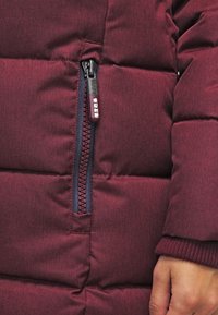 Bordowa kurtka puchowa z kieszenią na zamek, wykonana z tkaniny o fakturze, z poziomym pikowaniem oraz kontrastową taśmą zamka w ciemnym niebieskim kolorze.