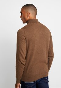 Pull-over tricoté marron avec un col haut et des manches longues, présentant une texture lisse. Le dos affiche une coupe ajustée et un ourlet côtelé.