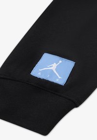 Μαύρο ύφασμα με μπλε ορθογώνιο έμπλαστρο που φέρει λευκό σιλουέτα λογότυπο και το κείμενο "AIR" σε μοντέρνα γραμματοσειρά στην μανσέτα.