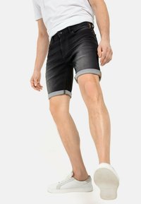 Schwarze Denim-Shorts mit aufgeschlagenen Säumen, die ein verblasstes Finish und einen dezenten gelben Akzent in der Nähe des Bundes aufweisen. Getragen mit weißen Sneakers.