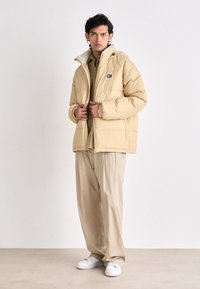Light beige puffer jacket with a soft, textured collar, zip closure, and elastic cuffs, paired with loose beige trousers and white sneakers.