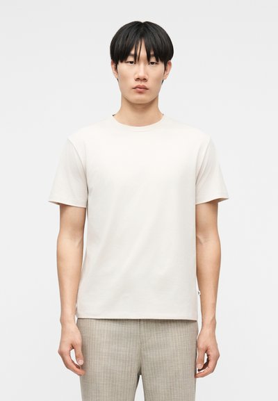 Jeune homme portant un t-shirt à manches courtes beige clair uni et un pantalon à carreaux gris clair, se tenant devant un fond blanc.