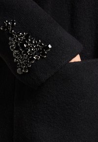 Zwart gebreid kledingstuk met een gestructureerd oppervlak, met een zak versierd met glanzende zwarte edelstenen in een driehoekig patroon.