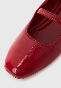 Zapato Mary Jane de charol rojo con punta cuadrada y una correa delgada sobre el empeine sobre un fondo blanco.