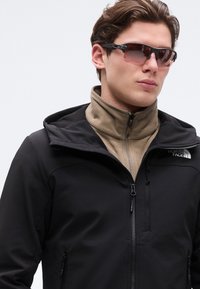 Jeune homme portant une veste noire North Face par-dessus une polaire beige et des lunettes de soleil sportives teintées foncées, regardant vers la droite sur un fond uni.