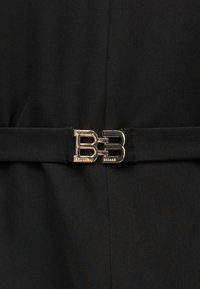 Αγκράφα διπλού Β σε χρυσό τόνο που κλείνει μια μαύρη υφασμάτινη ζώνη γύρω από μαύρα ρούχα.